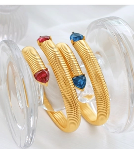 Detail of gold Tubogas bracelets featuring heart-shaped red and blue gemstones on a glass stand
