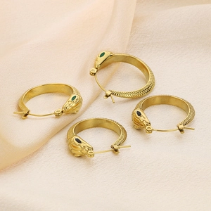 Golden snake-shaped hoop earrings with green and black enamel eyes on silk background