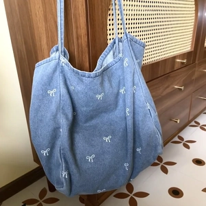 Large denim tote bag with white bow print and lace details on a vintage wood cabinet