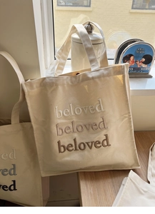 Detail of embroidered beige canvas tote bag showing the gradient beloved lettering