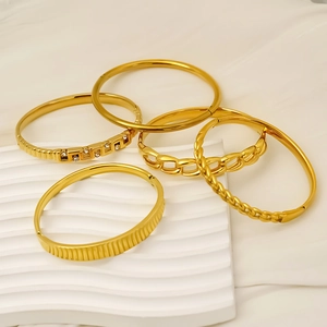 Five assorted gold stainless steel bracelets with various textures on a white wave board