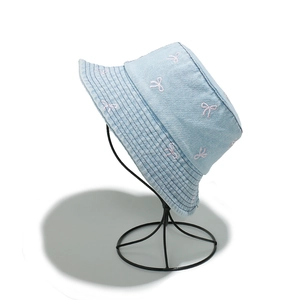 Detailed close-up of light blue denim bucket hat with pink bow embroidery on white stand