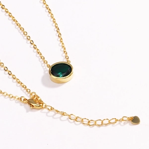 Detail view of a gold necklace with round emerald pendant and heart charm on the extender
