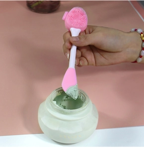 Dual-ended pink silicone face brush applying green mud mask from a white jar