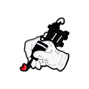 Cartoon enamel pin featuring a hand holding a black tattoo machine with a red heart icon