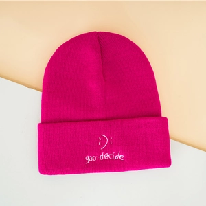Close-up of pink knit beanie featuring smiley face embroidery and you decide text