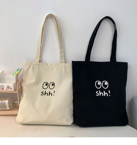 Detalle de bolsos tote de lona en blanco y negro con diseño de ojos y texto shh
