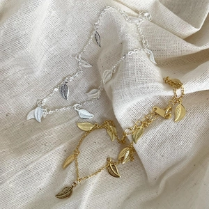 Detail of silver and gold anklets with leaf pendants on a soft beige fabric background