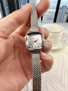 Close-up of a silver square watch with mesh band held against a blurry city background