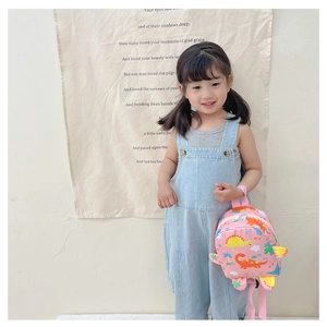 Toddler girl wearing pink dinosaur backpack with blue denim overalls in a lifestyle setting