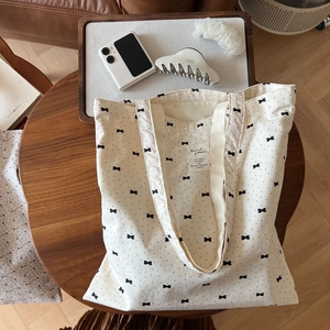 Cream canvas tote bag with black floral bow patterns and shoulder straps on a wooden table