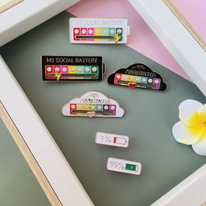 Detail view of enamel social battery pins and mood tracker badges on a decorative background