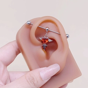 Dettaglio del piercing industriale argento con gemma a cuore rossa e ali sull'orecchio