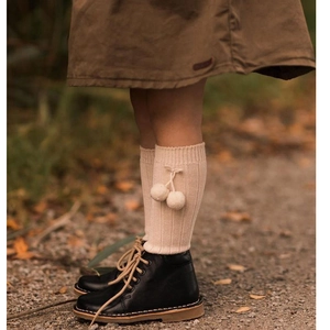 Detail of cream knee-high baby socks with pom-poms paired with black lace-up boots