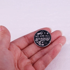 Close-up of circular We Are All Stardust enamel pin held in hand for size comparison