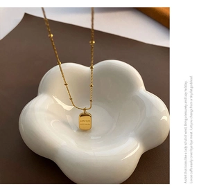 Detailed view of a gold square charm pendant with bead chain on a ceramic plate