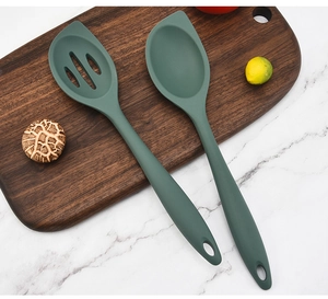Green silicone spoon and slotted spatula set on a dark wood board with fresh ingredients