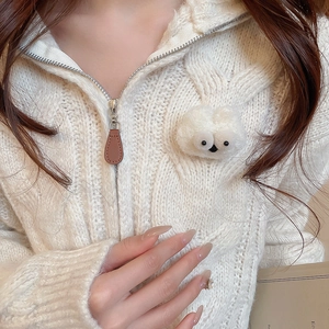 Detailed close-up of white cable knit sweater with plush doll brooch and leather zipper pull