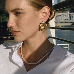 Model trägt goldene Blumen-Ohrstecker und Perlenhalskette am Wasser im Freien