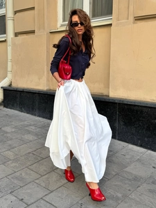 Full-length street shot of a long white pleated skirt with a navy blue cardigan and red heels
