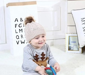 Close-up of a beige cable-knit baby hat with faux fur pom pom on a baby model