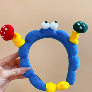 Detailaufnahme eines blauen Plüsch-Haarreifs mit großen Augen und bunten Pilz-Applikationen