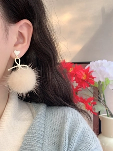 Close-up of white heart studs with ribbon and soft beige pompom earrings on a model