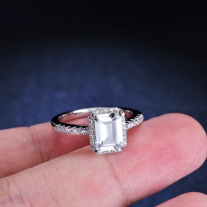 Emerald cut moissanite ring with halo setting on finger over blue background