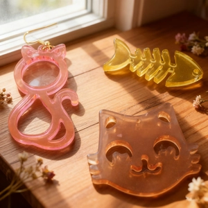 Handmade resin cat and fishbone charms with gold hooks on a sunlit wooden background