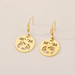 Detailed close-up of gold MOM heart earrings with baby footprints on a beige surface
