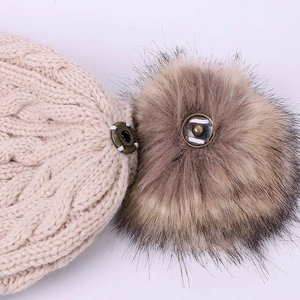 Close-up of beige cable knit hat with metal snap button and brown faux fur pom-pom