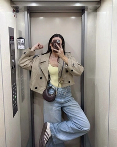 Short double-breasted khaki trench coat styled with blue wide-leg jeans in elevator selfie