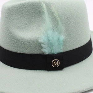 Détail d'un chapeau fedora vert menthe avec ruban noir, plumes et logo doré en métal