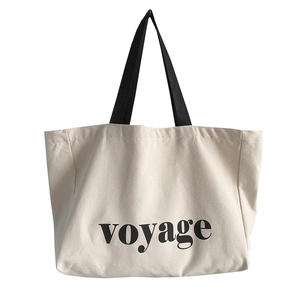 Large beige canvas tote bag with black handles and voyage lettering print for travel