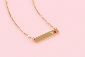 Close-up of golden bar pendant with engraved heart and red gemstone on a pink background