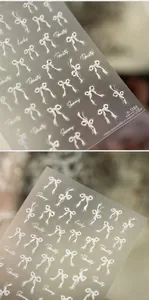 Makro-Detail von weißen Schleifen-Nagelstickern mit Perlmuttglanz und Schrift m-1266