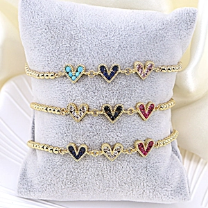 Three gold beaded bracelets with colorful zircon heart charms on gray velvet pillow
