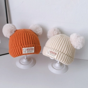 Cute knitted baby beanies in orange and cream with twin white fluffy pom-poms on a stand