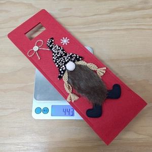 Close-up of a red felt gnome wine bottle cover with snowflake decor on a weight scale