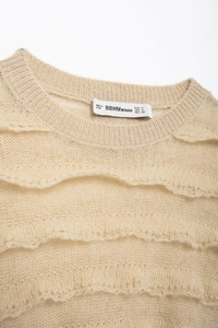 Detail view of beige knit sweater showing layered ruffle trim and delicate texture