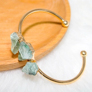 Detail of gold-plated open bracelet featuring raw green gemstones on wooden tray