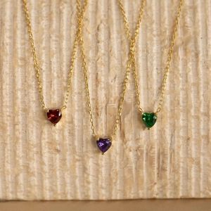 Three gold heart birthstone necklaces in red, purple, and green on textured stone background