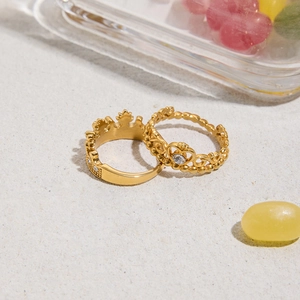 Dettaglio di anelli corona e cuore in acciaio dorato 18K con strass su sabbia bianca