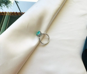 Detailed view of emerald cut teal gemstone ring set on silver band over silk cloth