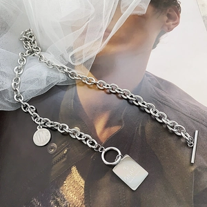 Detalle de collar de acero plateado con cierre OT y colgante grabado sobre malla blanca
