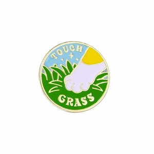 Detail of round metal enamel pin featuring a cat paw reaching for grass with Touch Grass text