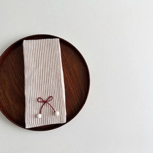 Detail of beige ribbed knit scarf featuring deep red bow and white fuzzy pom-poms