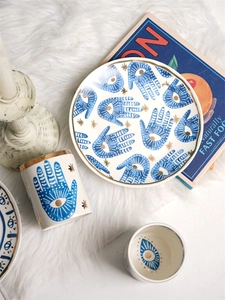 Flat lay of ceramic Hamsa hand plate and cup with gold stars on a plush white background