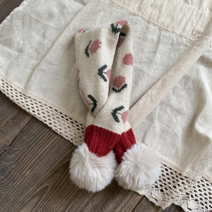 Detail of beige knit scarf featuring pink floral patterns and white faux fur pom-poms