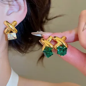 Crossed gold earrings with square emerald crystal pendants worn by a model